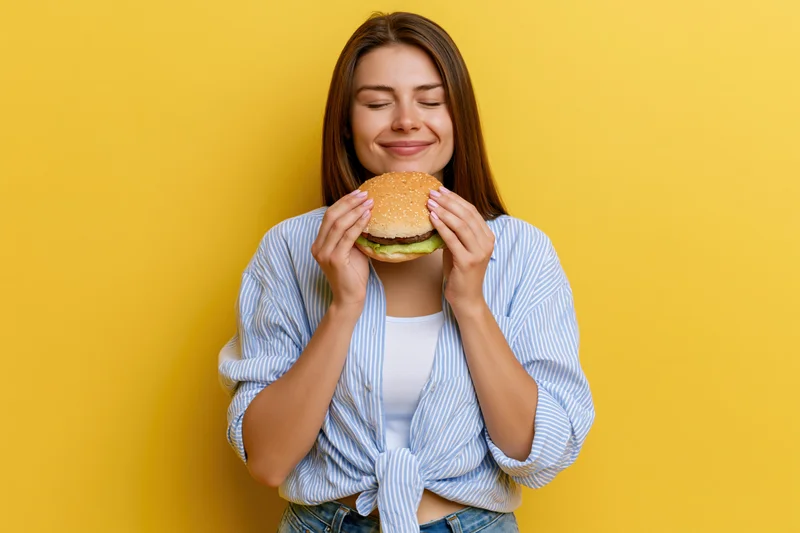 Young woman holding a burger with eyes closed and a satisfied smile against yellow background
