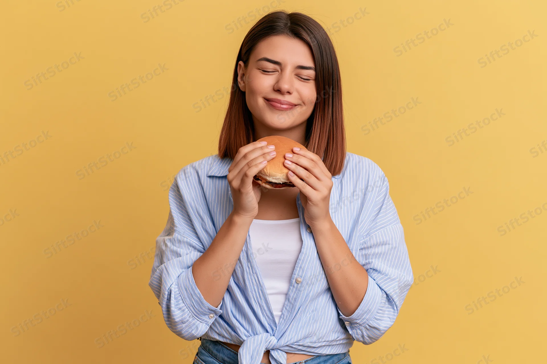 Woman holding a burger with eyes closed and smiling against yellow background