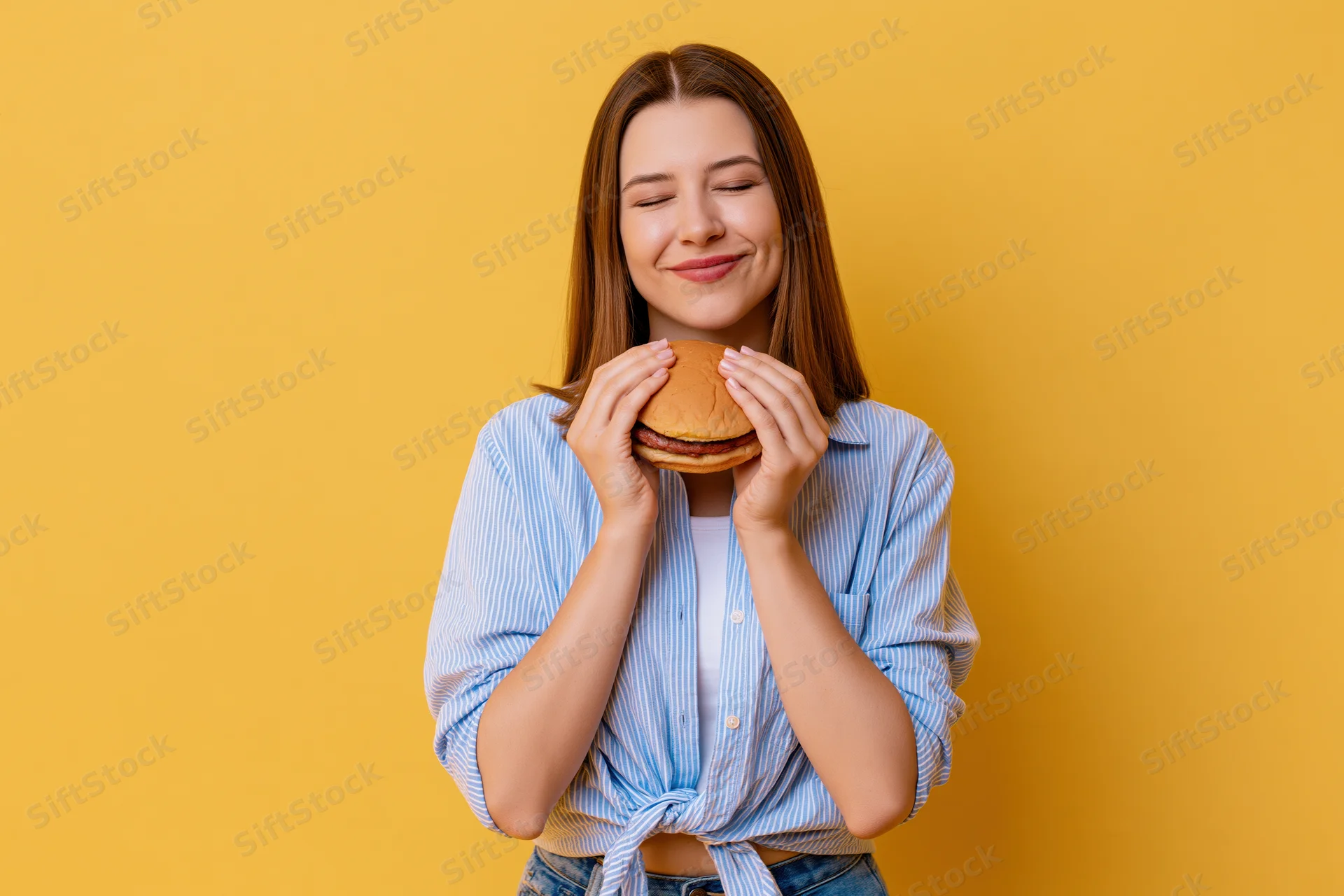 Young woman holding a burger with eyes closed and smiling against yellow background