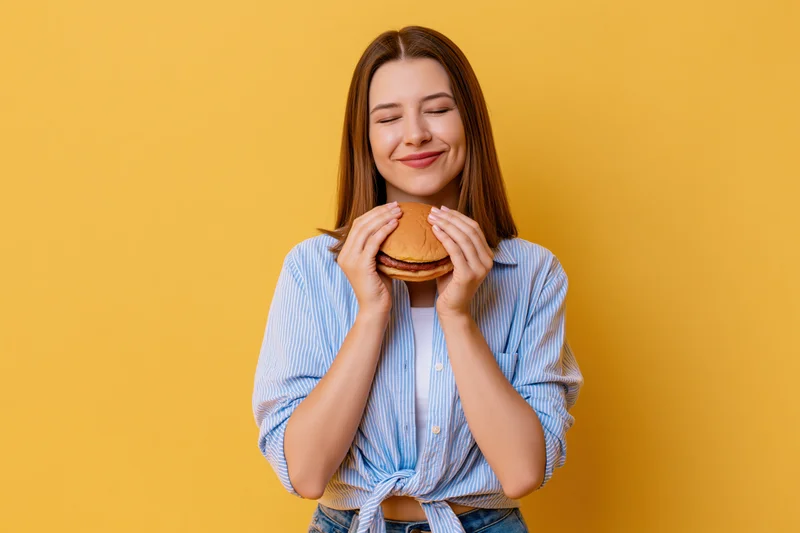 Young woman holding a burger with eyes closed and smiling against yellow background