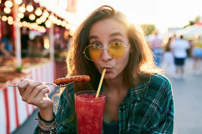 Young woman sipping red drink and eating sausage on a fork at a summer festival