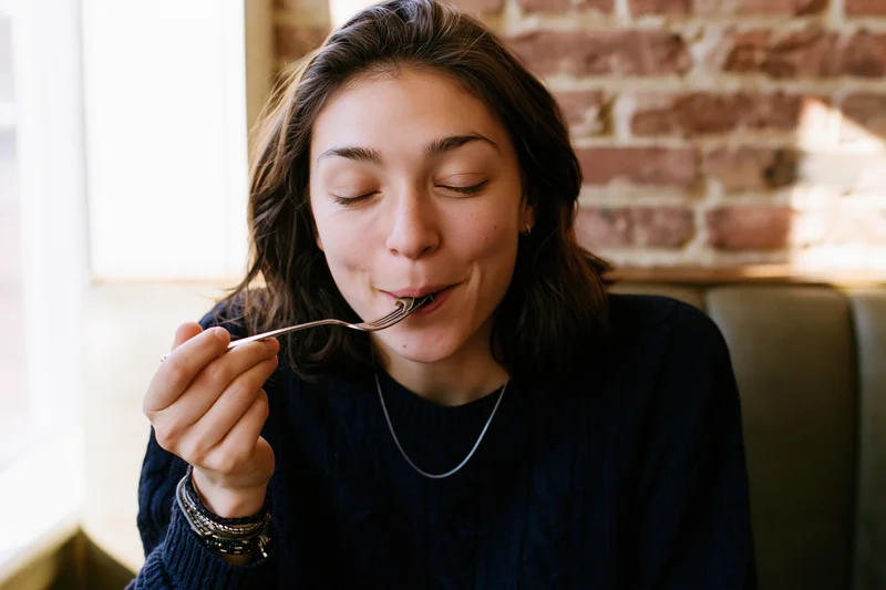 Woman eating with fork, eyes closed in pleasure, seated in restaurant booth near window