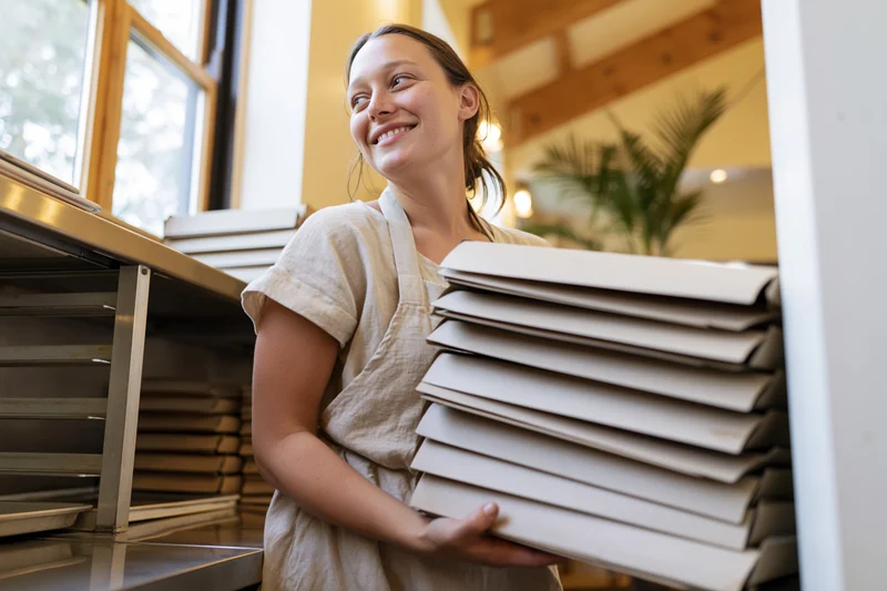 Young woman in apron smiling while holding stack of bakery boxes in kitchen