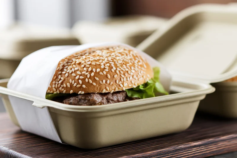 Beef burger with sesame seed bun and lettuce in open takeout container on wooden surface