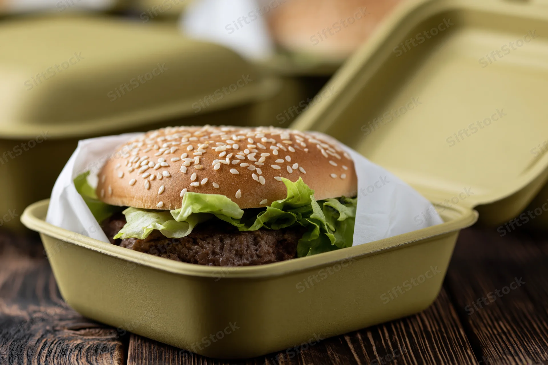 Beef burger with sesame seed bun and lettuce in a green takeout container on a wooden table