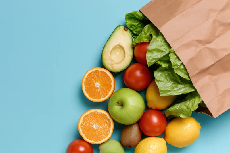 Brown paper grocery bag tipped over spilling fresh fruits and vegetables on blue surface