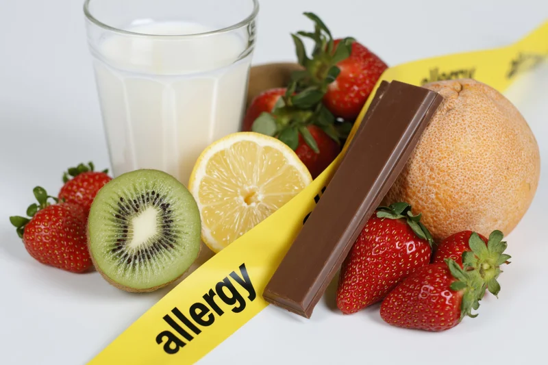 Various fresh fruits and a glass of milk arranged with a yellow allergy warning tape on a white surface