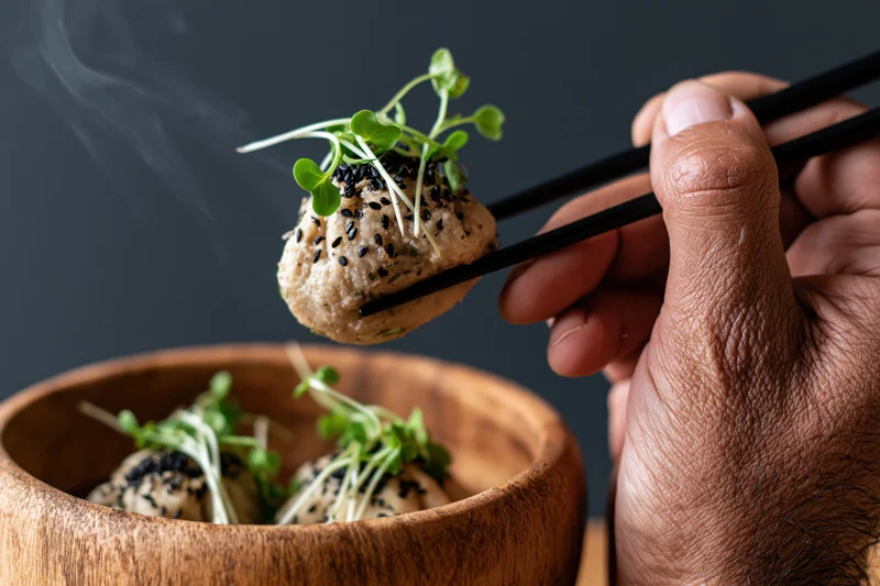 Hand holding chopsticks lifting a steaming dumpling from a wooden bowl