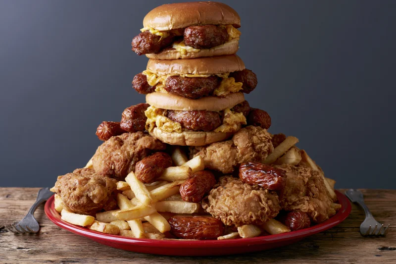 Towering stack of sausage burgers and fried chicken with fries on a red plate on a wooden table
