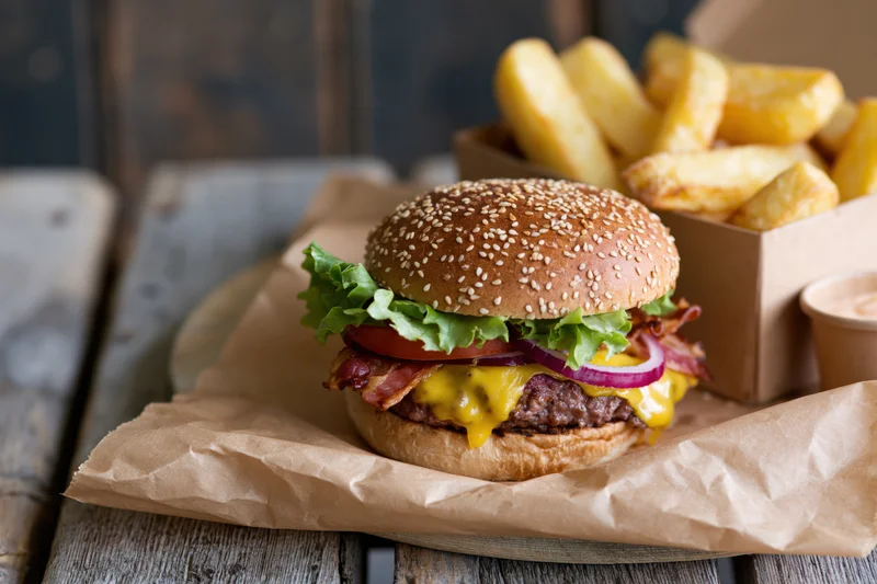 Cheeseburger with lettuce tomato red onion bacon and melted cheese on sesame bun served with french fries on paper wrapper on wooden table