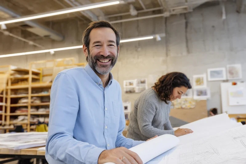 Smiling architect holding blueprint in modern office workspace with colleague working in background