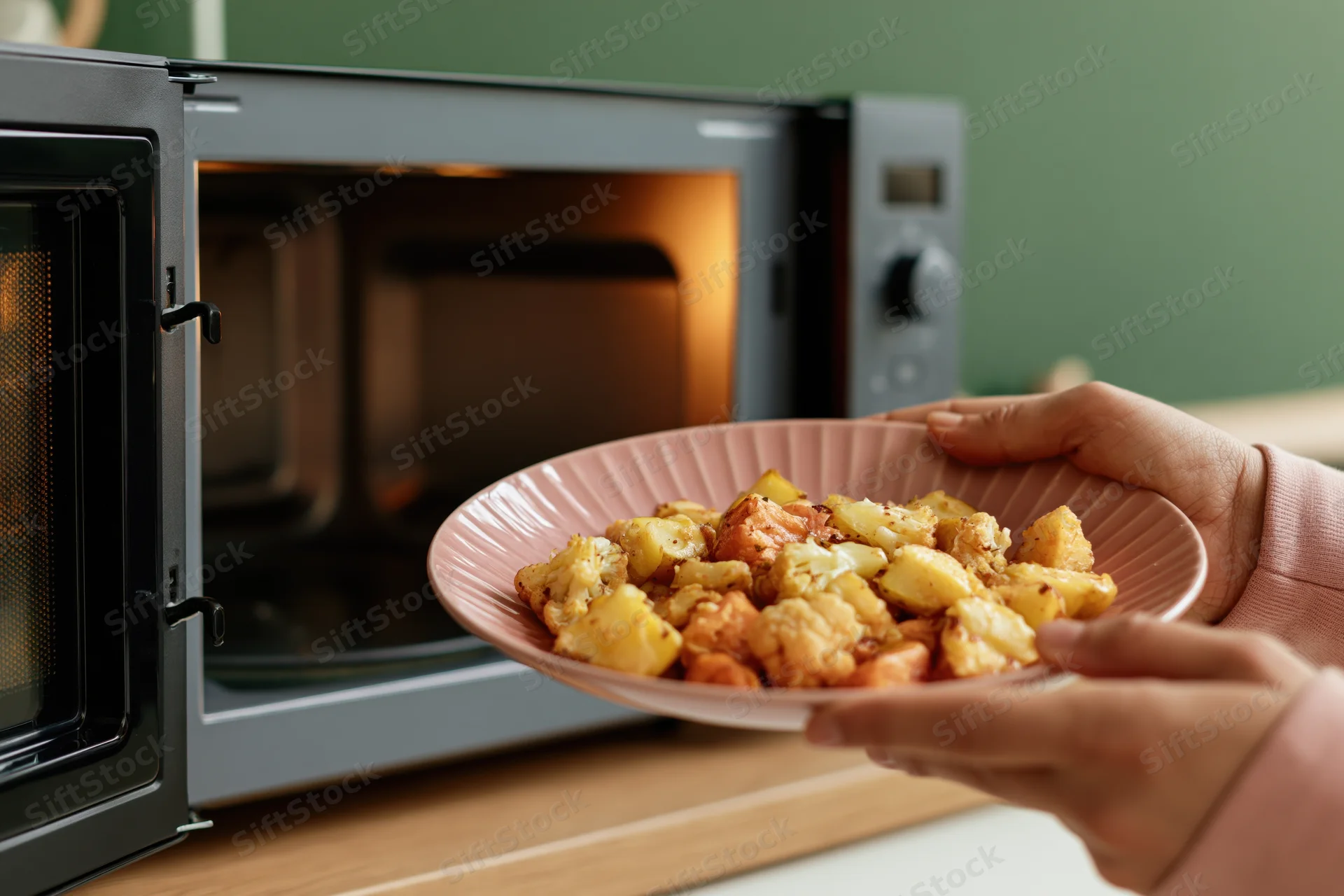 Woman holding a pink plate of roasted vegetables and potatoes in front of an open microwave on a kitchen countertop