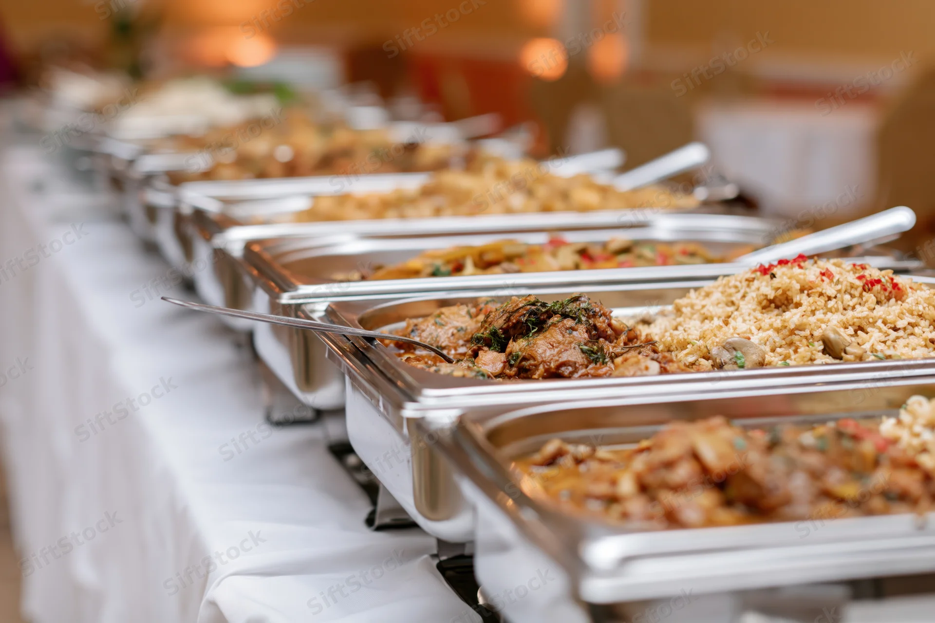 Row of stainless steel chafing dishes filled with hot food on a white tablecloth at an event