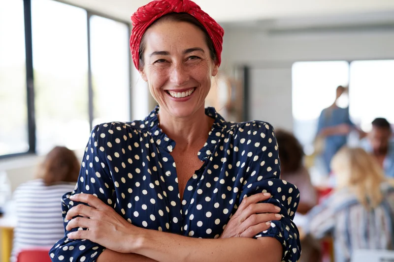 Smiling woman with arms crossed wearing red headscarf and polka dot blouse in cafe