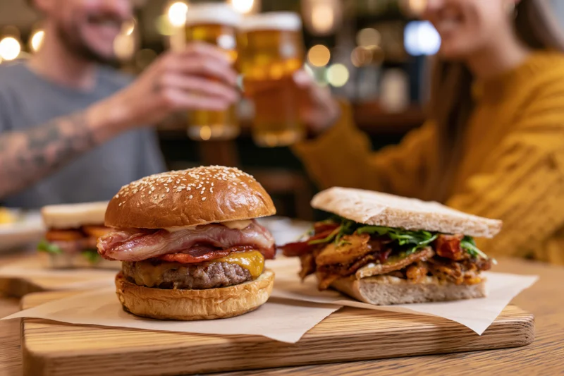 Stacked brioche burger with bacon and cheese on wooden board beside a sandwich, with couple toasting beer glasses in background