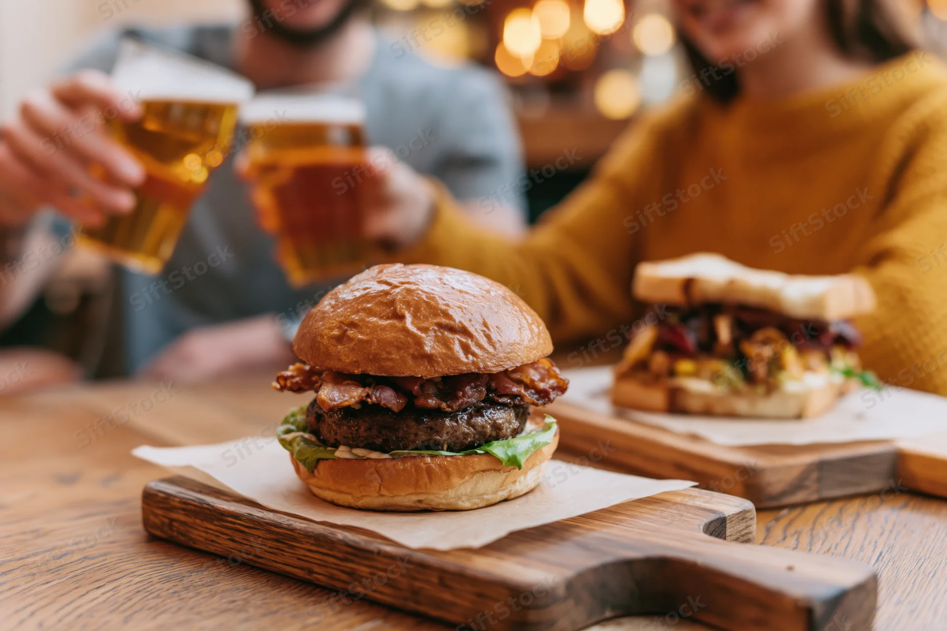 Burger with bacon and lettuce on wooden board with couple toasting beer in background
