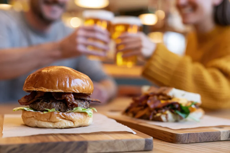 Stacked brioche burger on wooden serving board with couple toasting beer in background