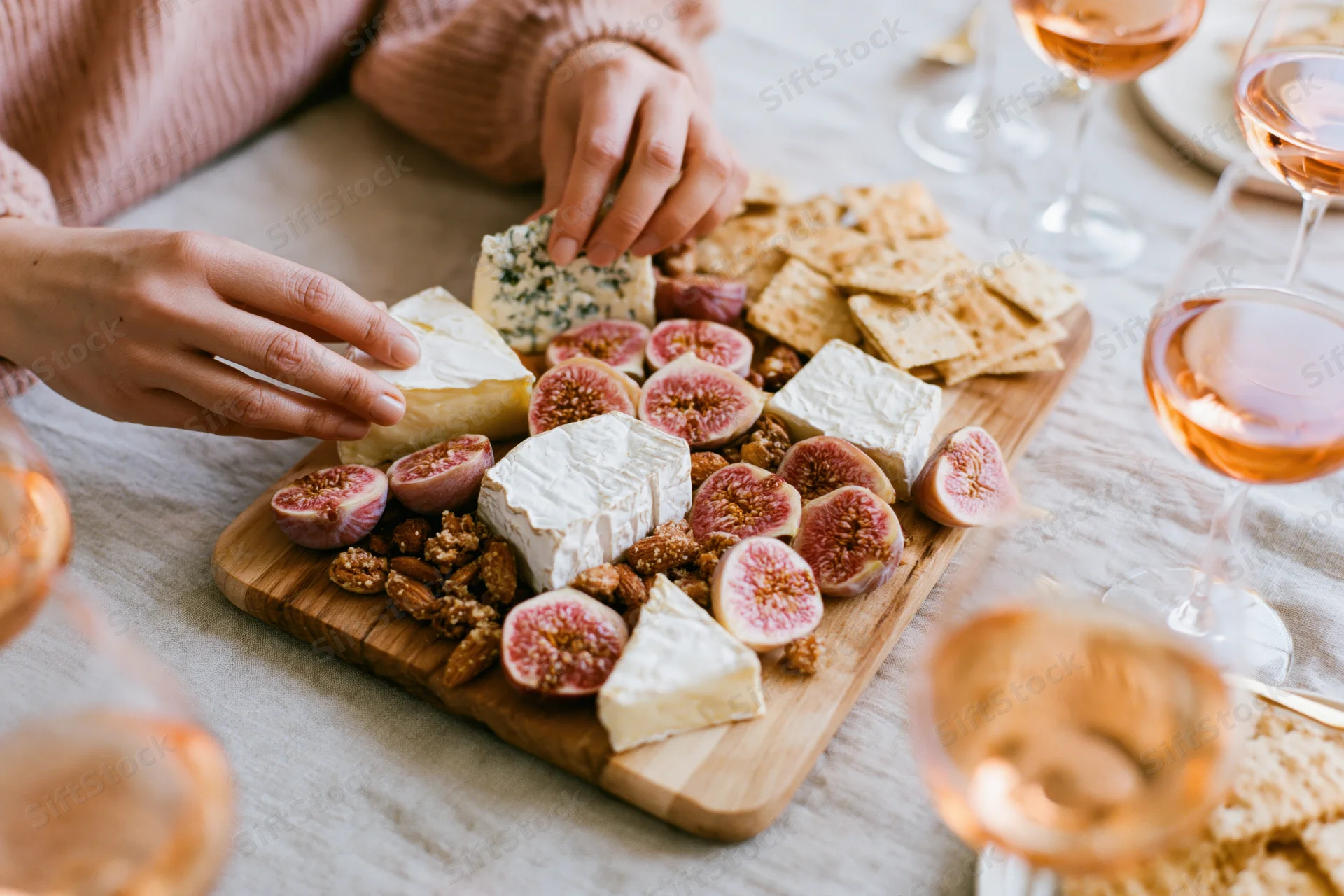 Woman's hands arranging cheese and figs on a wooden charcuterie board with wine glasses around