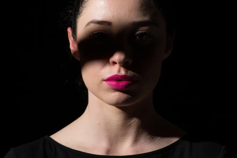 Young woman with dark hair pulled back in a dramatic studio portrait with pink lipstick and shadowed lighting