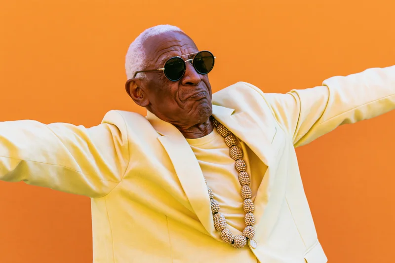 Senior man in yellow suit with sunglasses and beads posing with arms outstretched against orange background