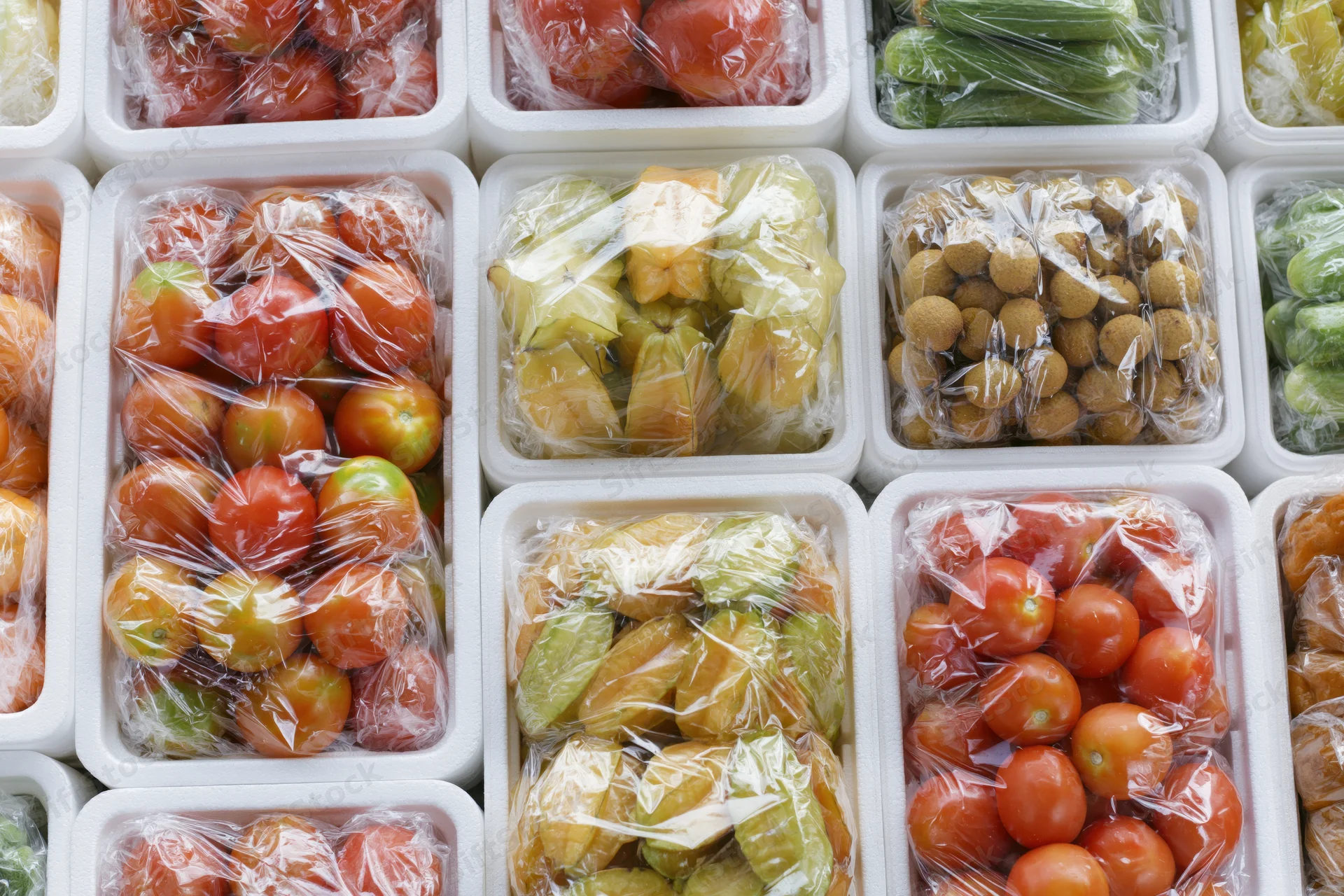 Assorted tropical fruits and vegetables in plastic-wrapped styrofoam containers arranged in a grid on a shelf