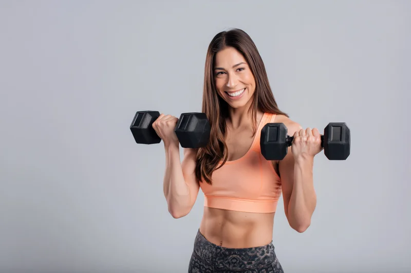 Woman lifting dumbbells in fitness attire smiling at camera in studio