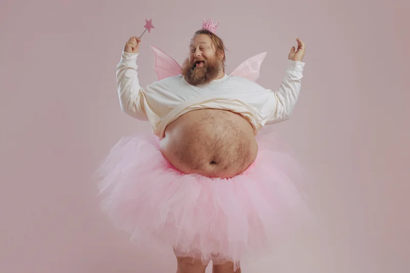 Bearded man in fairy costume with tutu and wand posing joyfully against pink background
