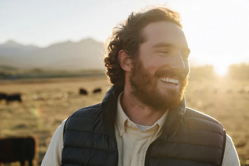 Smiling bearded man in puffer vest standing in a pasture with cattle at sunset