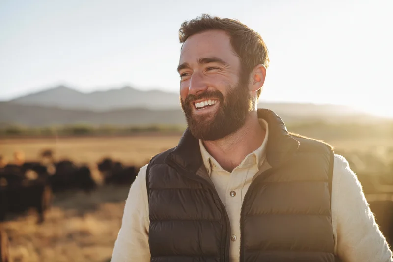 Smiling bearded man in puffer vest standing in a field with cattle at sunset