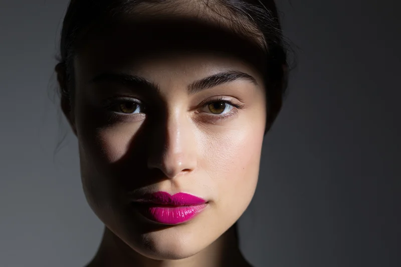 Closeup portrait of young woman with dark hair pulled back and bright pink lipstick under dramatic studio lighting