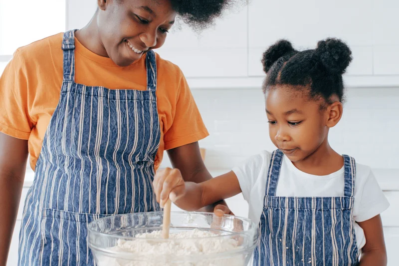 Black mother and young daughter mixing flour in a bowl in a bright kitchen
