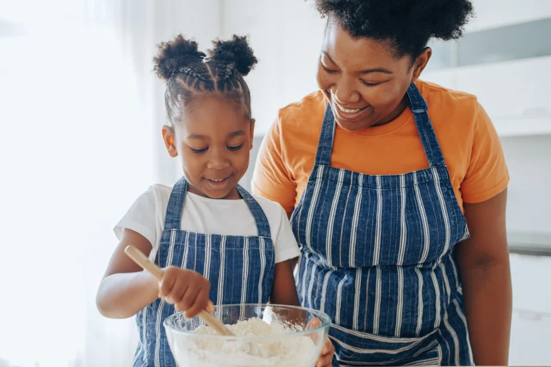 Black mother and young daughter mixing flour in a glass bowl while baking together in a bright kitchen