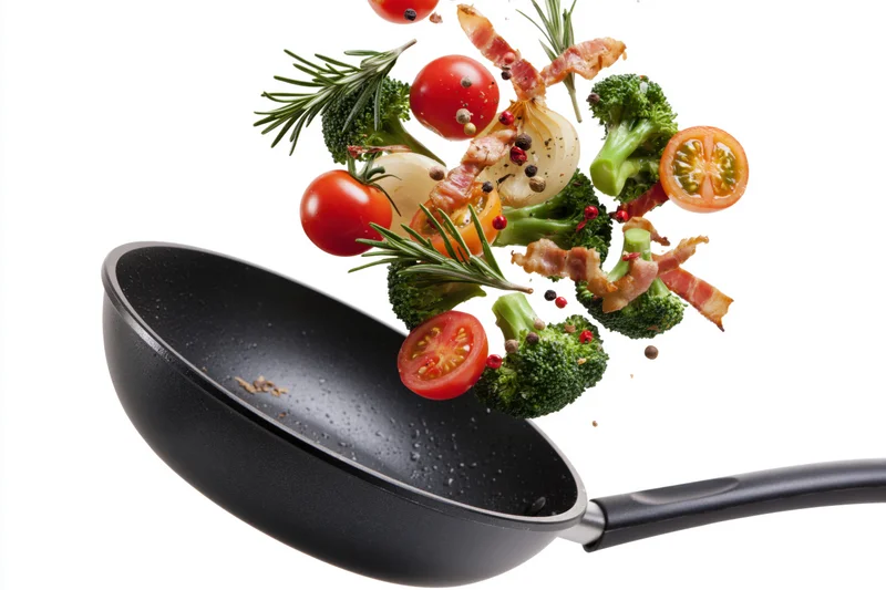 Black nonstick frying pan tilted mid-toss with vegetables and bacon flying out against white background