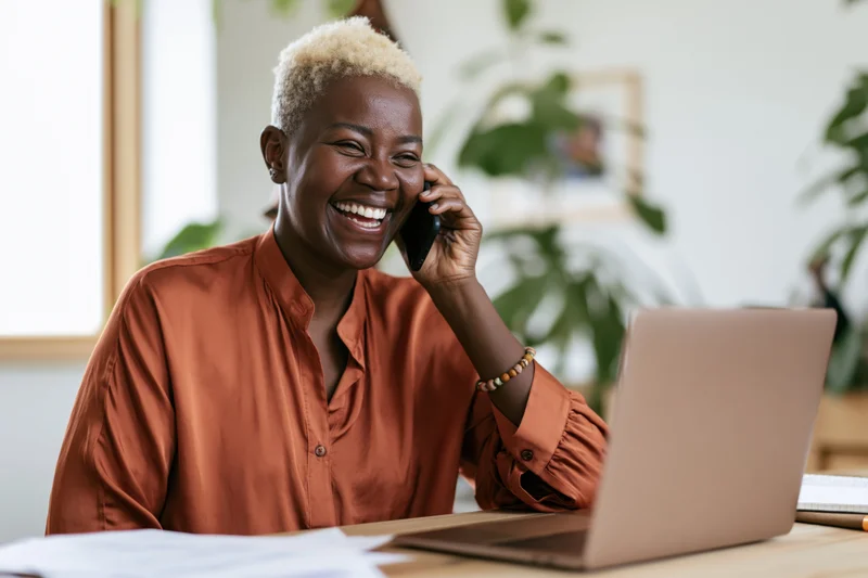 Black woman laughing while talking on phone at desk with laptop and papers