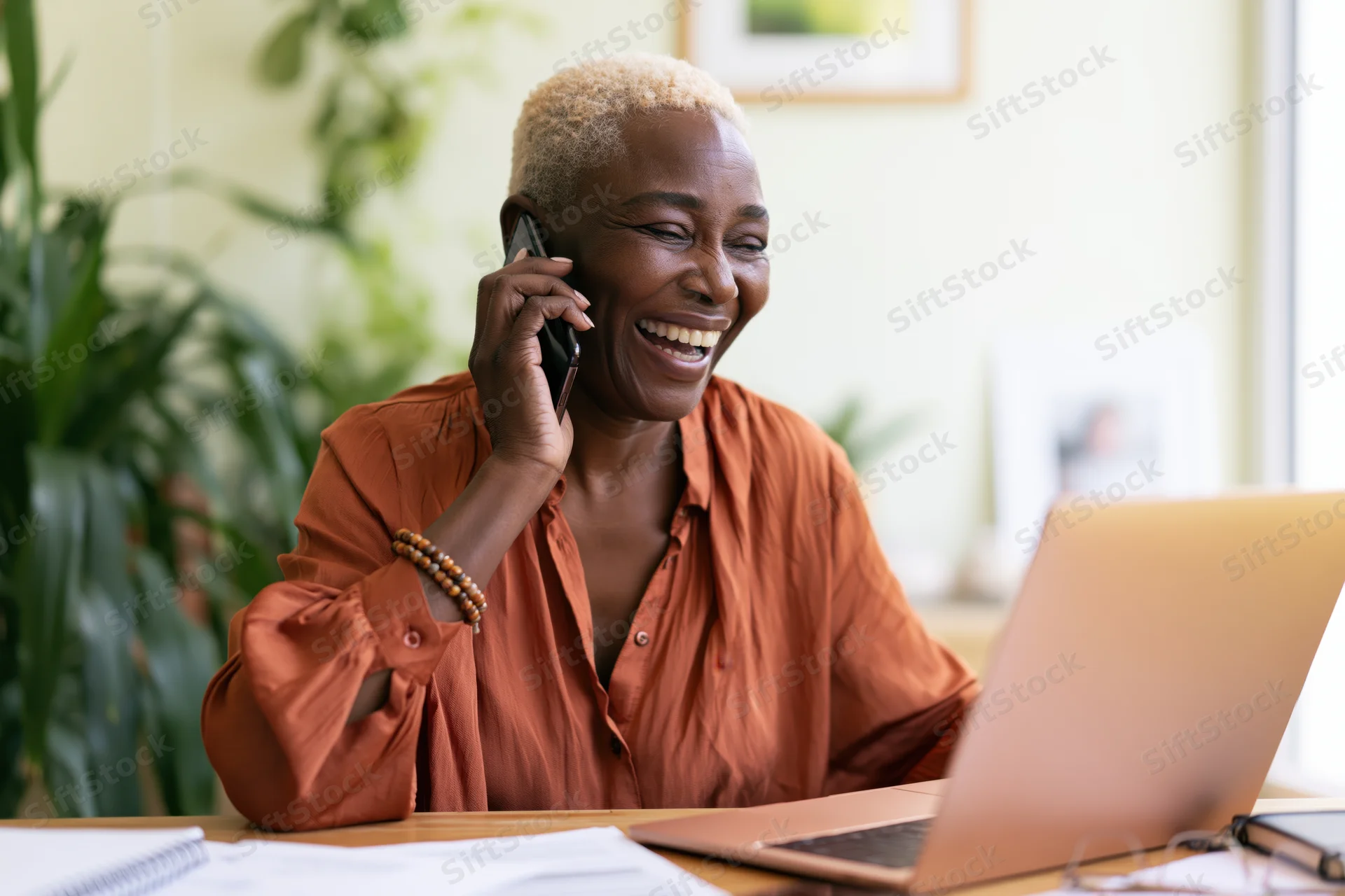 Black woman laughing while talking on phone at desk with laptop in home office