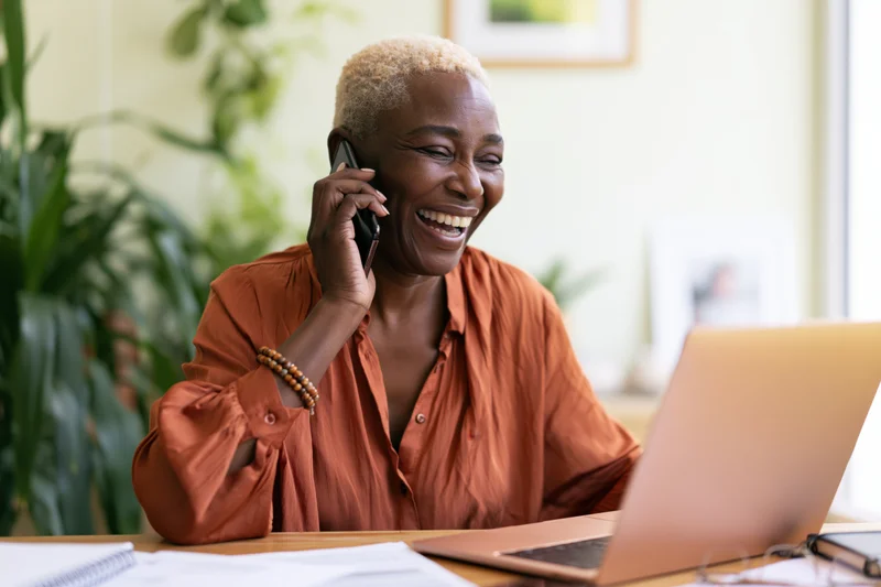Black woman laughing while talking on phone at desk with laptop in home office