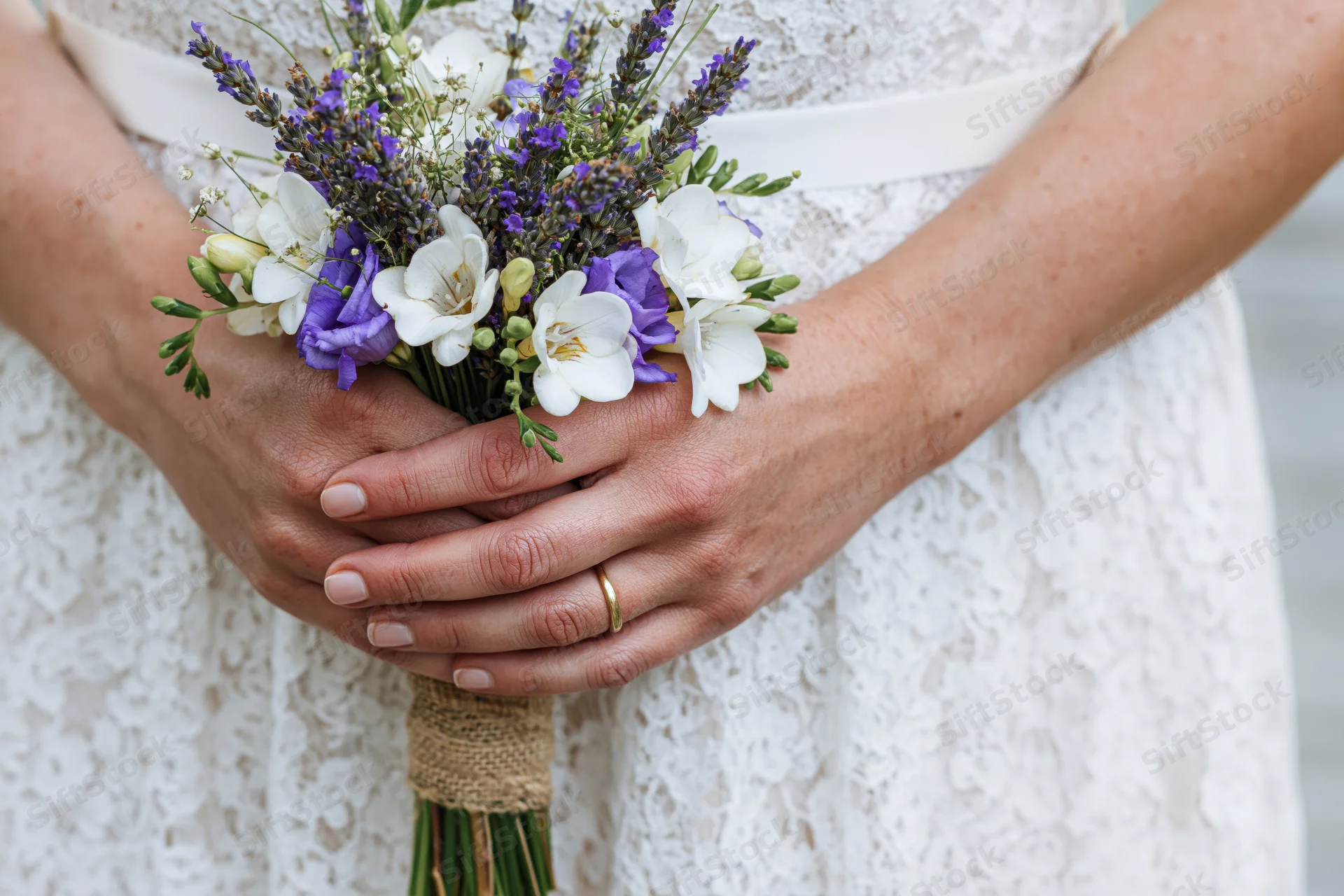 Bride holding rustic wedding bouquet with lavender and white flowers, hands clasped around stems, lace dress visible