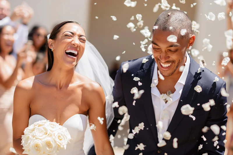 Bride and groom laughing as white petals fall around them at outdoor wedding ceremony