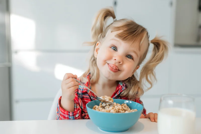 Toddler girl eating cereal with spoon at kitchen table, smiling playfully
