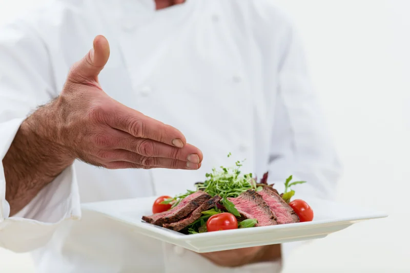 Chef presenting a plated steak salad with tomatoes and greens on a white plate