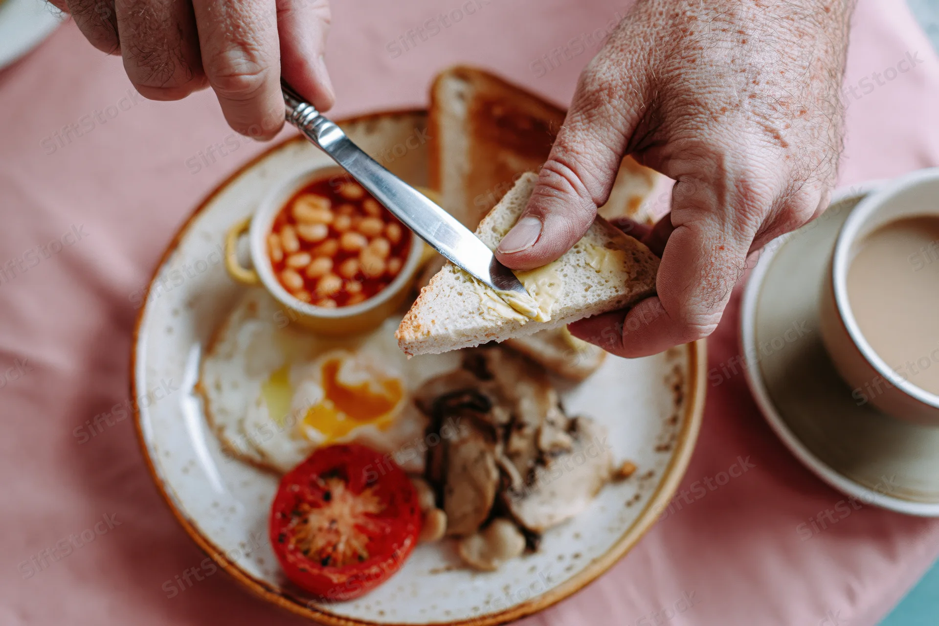 Man buttering a triangle of toast with a knife over a breakfast plate with eggs, beans, tomato and mushrooms, beside a cup of coffee on a pink tablecloth