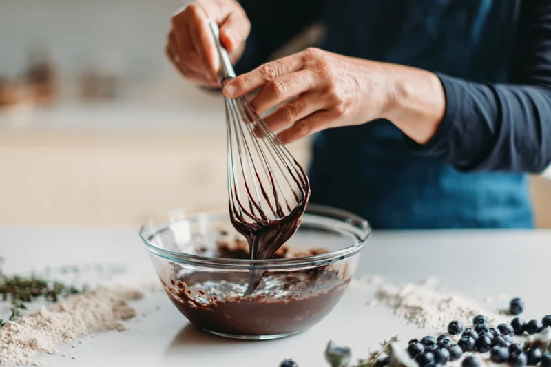 Woman mixing chocolate batter with a whisk in a glass bowl on a kitchen counter surrounded by flour, blueberries and thyme