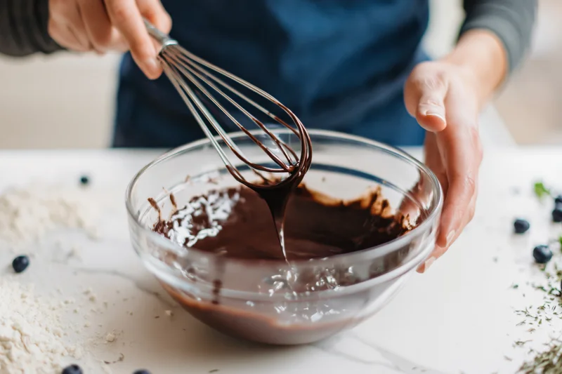 Woman mixing chocolate batter with a metal whisk in a glass bowl on a marble countertop