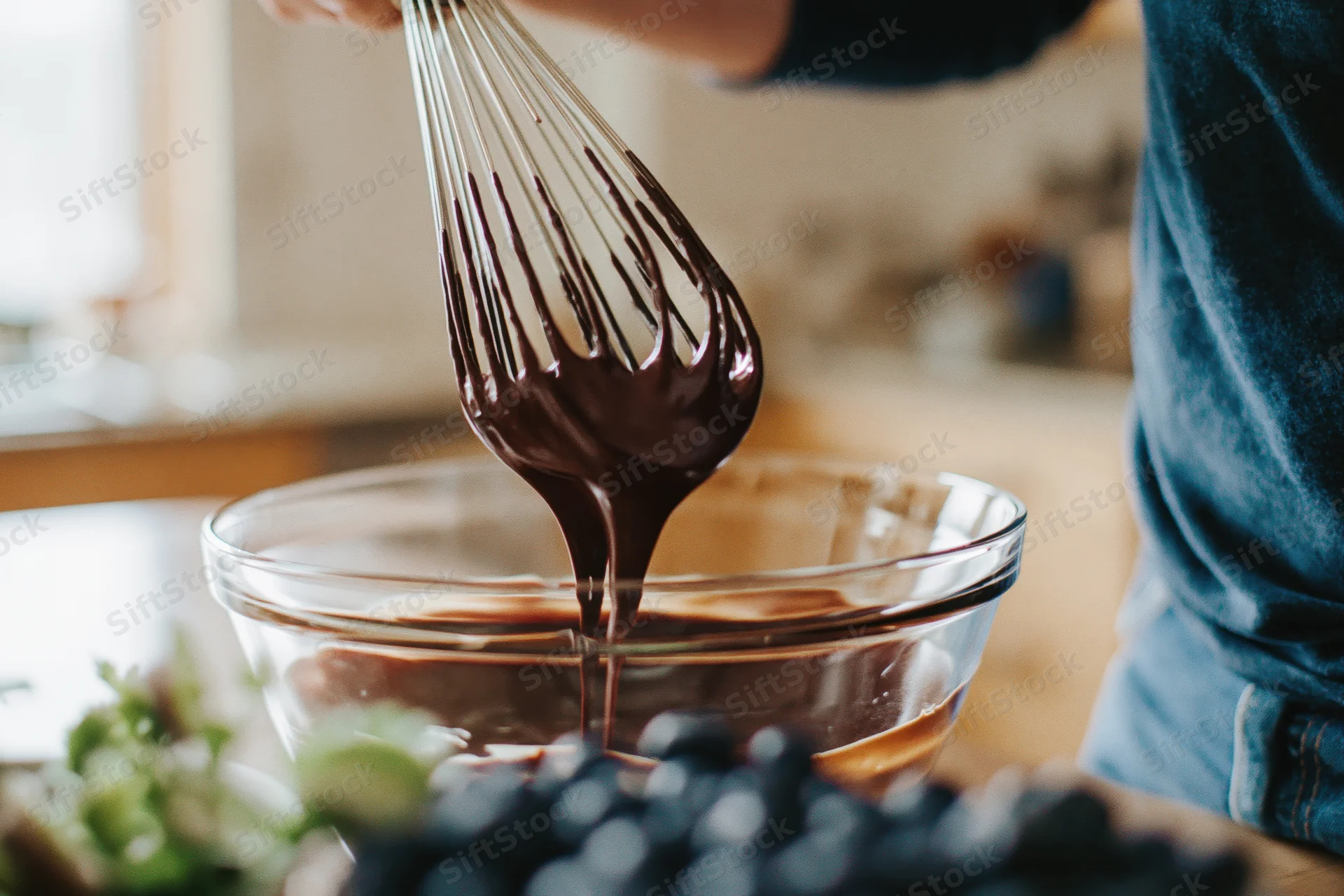 Hands lifting a wire whisk dripping thick melted chocolate over a glass bowl in a kitchen