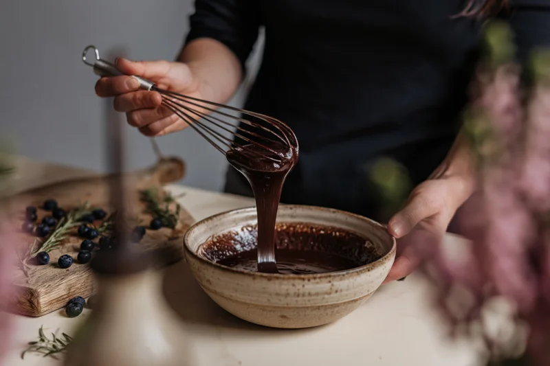 Woman lifting a whisk from a bowl of chocolate sauce in a kitchen setting