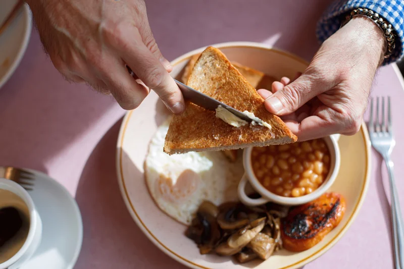 Man buttering toast with knife over full english breakfast plate on pink table