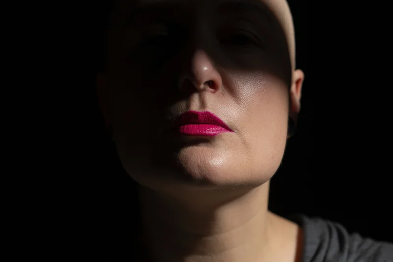 Close-up portrait of a bald woman with pink lipstick against a dark background