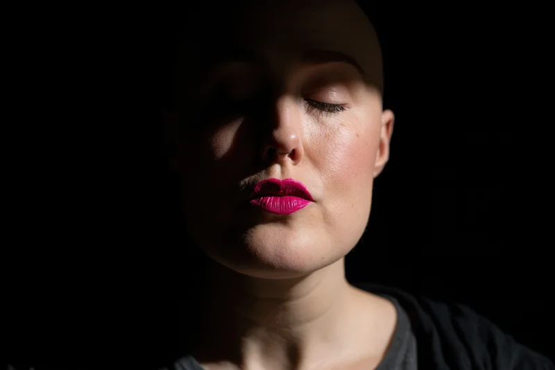 Close-up portrait of a bald woman with pink lipstick against a dark background