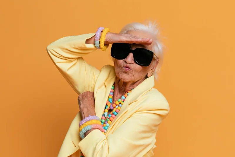 Elderly woman in yellow jacket doing dab pose with hand over eyes against orange background