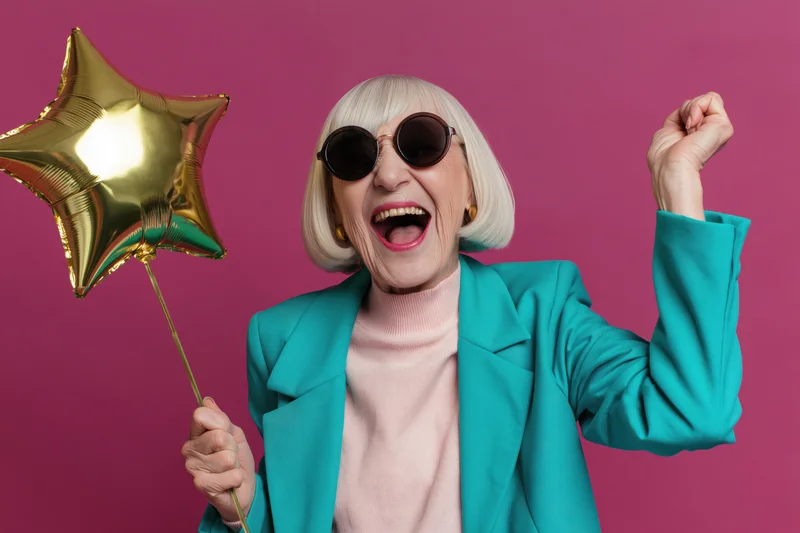 Elderly woman laughing widely while holding a gold star balloon and raising her fist against a pink background